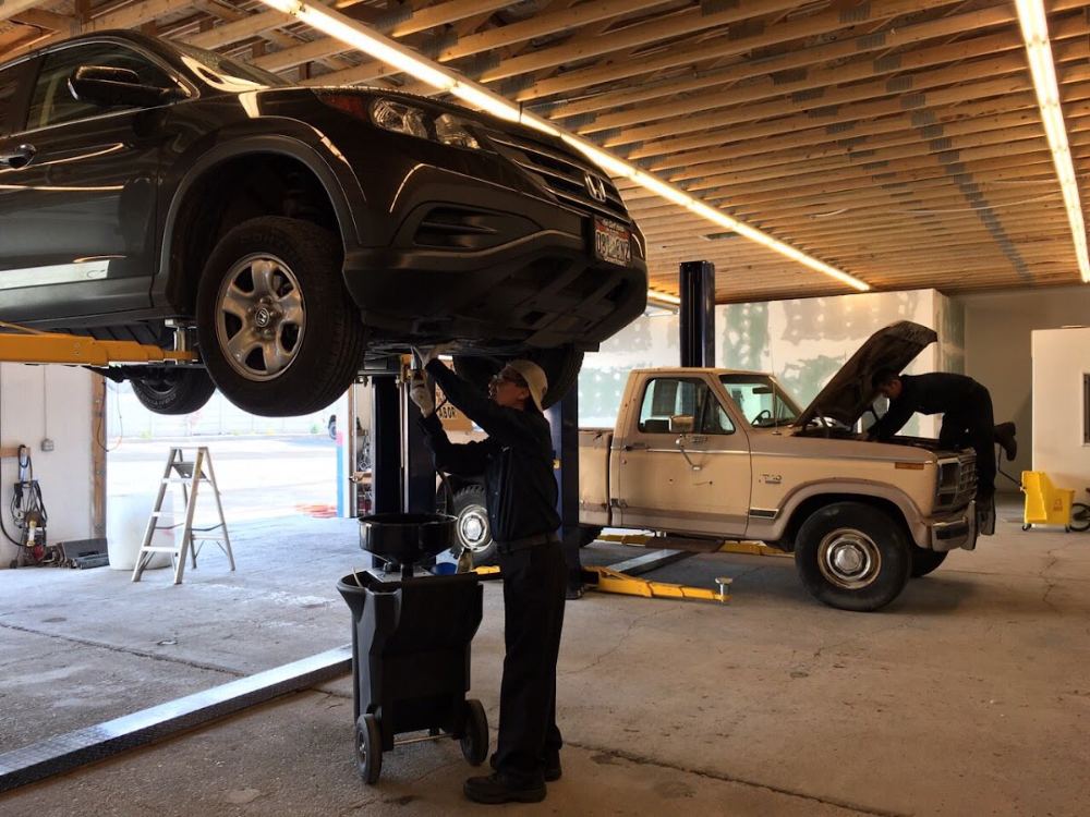 A mechanic draining oil under an SUV on the lift at Unitech Automotive, with an old Ford pickup in the next bay